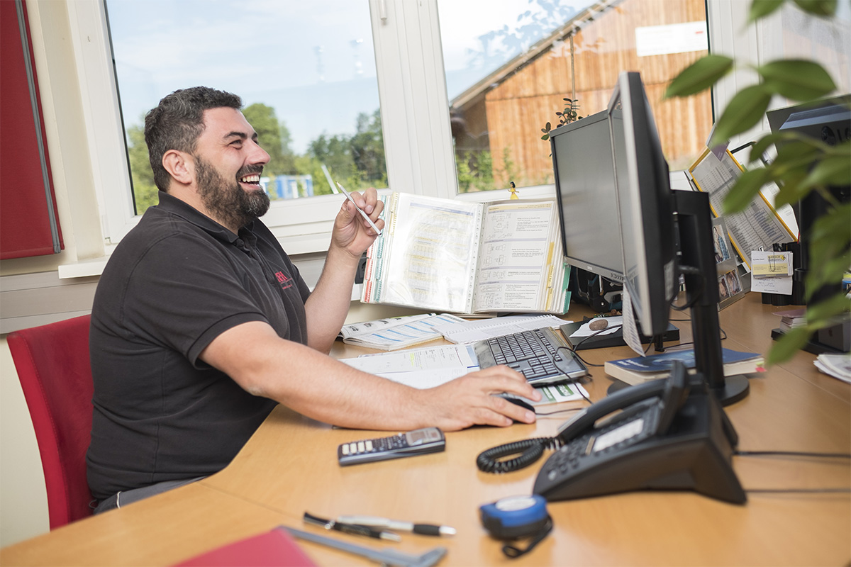 Kontakt. Ein Mitarbeiter sitzt sehr vergn&uuml;gt lachend vor einem Bildschirm an einem Schreibtisch.