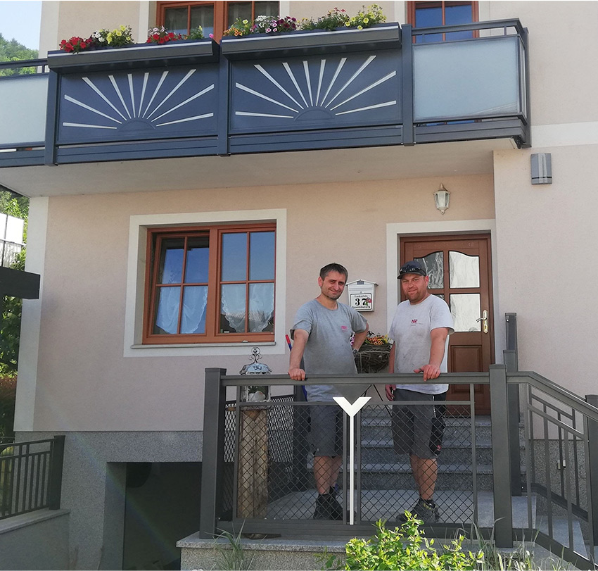 Vor einem Haus stehen zwei Männer unter einem Balkon mit Sichtschutzplatten mit geometrischem Muster und stützen sich auf das Eingangsgeländer.